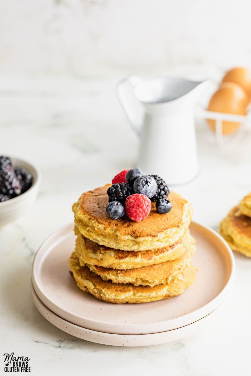 A stack of almond flour pancakes with berries on top. A can of milk and eggs in the background.