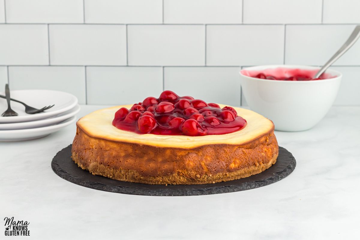 Gluten-free cheesecake with cherry topping on a cake dish, with a stack of plates and a bowl of cherry topping in the background.