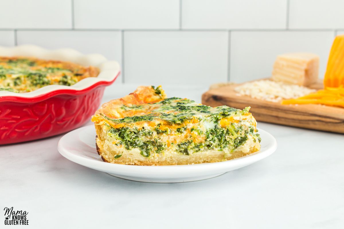 A slice of gluten-free spinach quiche on a plate with some quiche and cheese in the background.