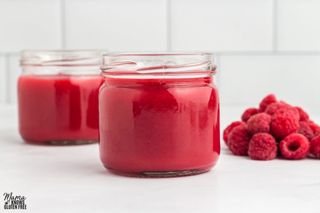 Two jars of  Raspberry coulis and some fresh raspberries on the side.