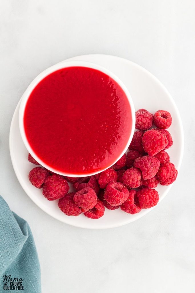 Raspberry coulis in a cup on a plate with some fresh raspberries.