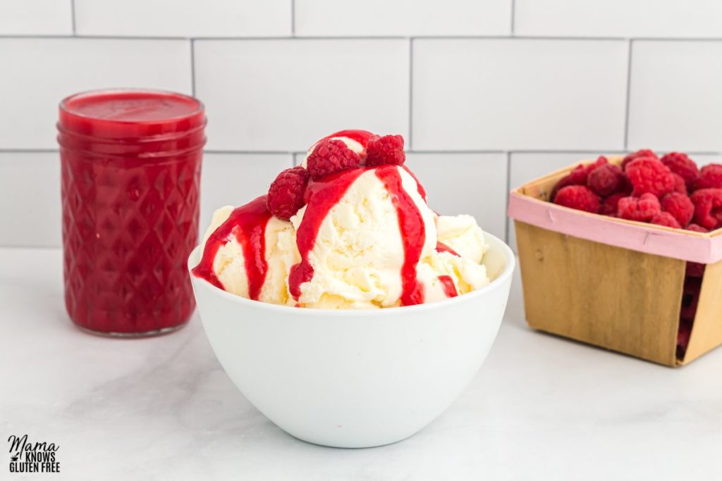 A bowl of ice cream topped with  Raspberry coulis and fresh raspberries. A jar of Raspberry coulis and some fresh raspberries on the side.