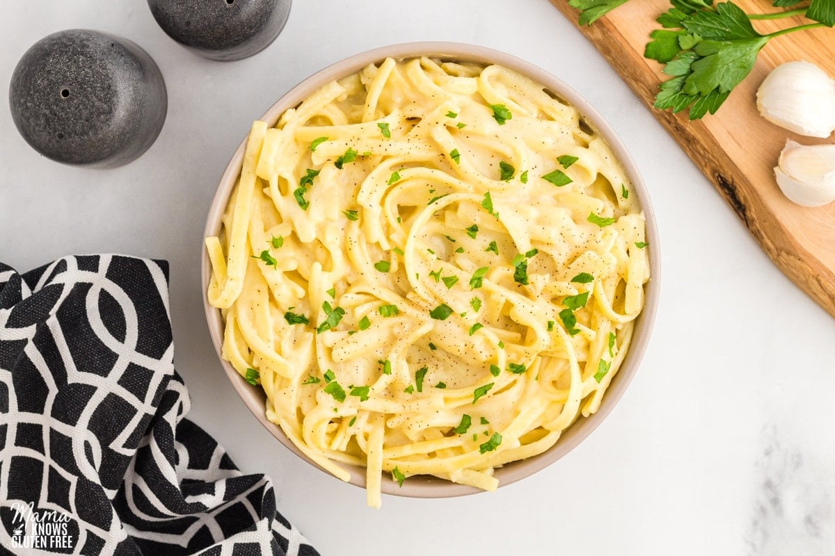 dairy-free Alfredo sauce over pasta in a cream bowl with a black napkin, salt and pepper shakers, and cutting board in the background.