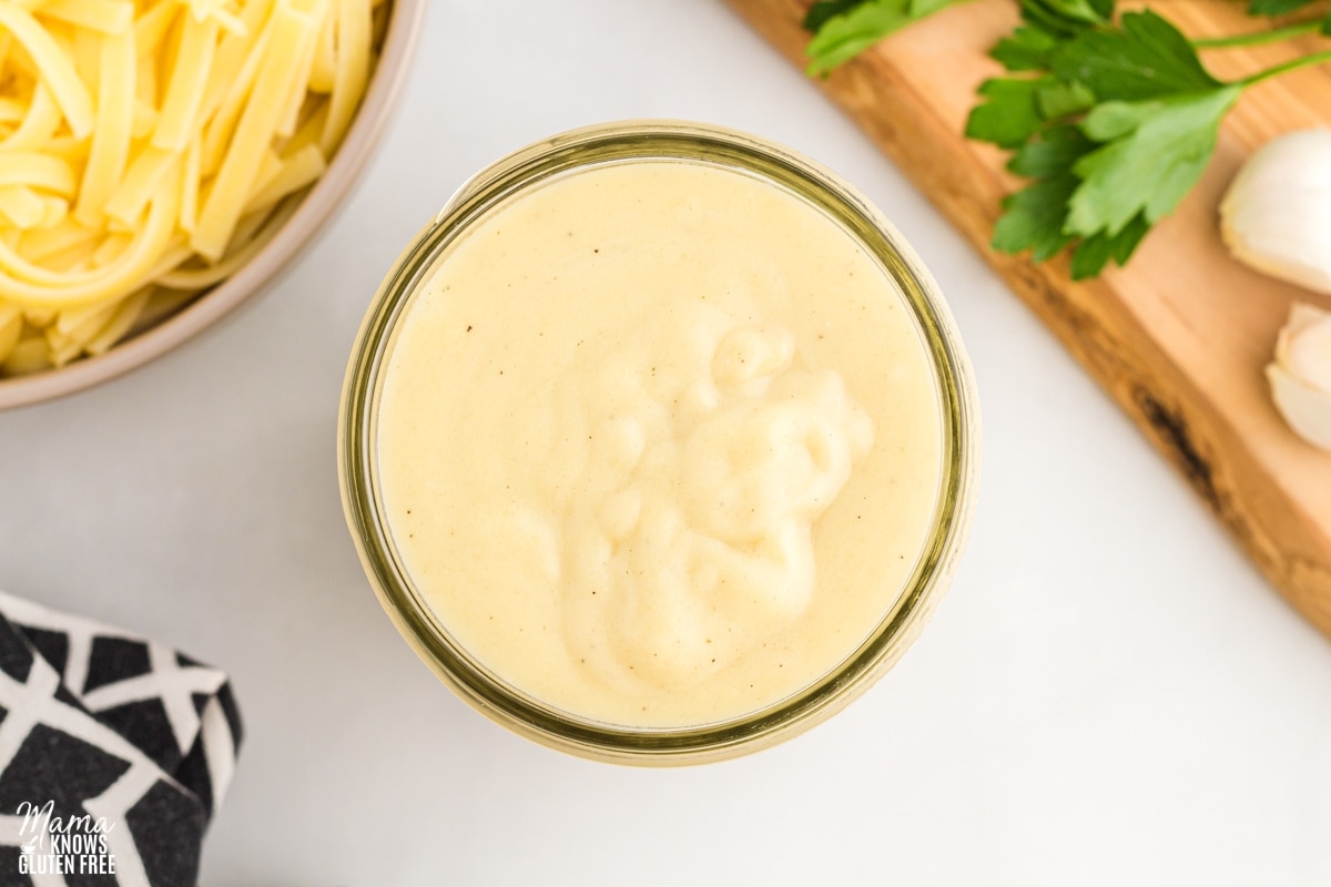 dairy-free alfredo sauce in a glass jar with pasta, black napkin, and cutting board in the background