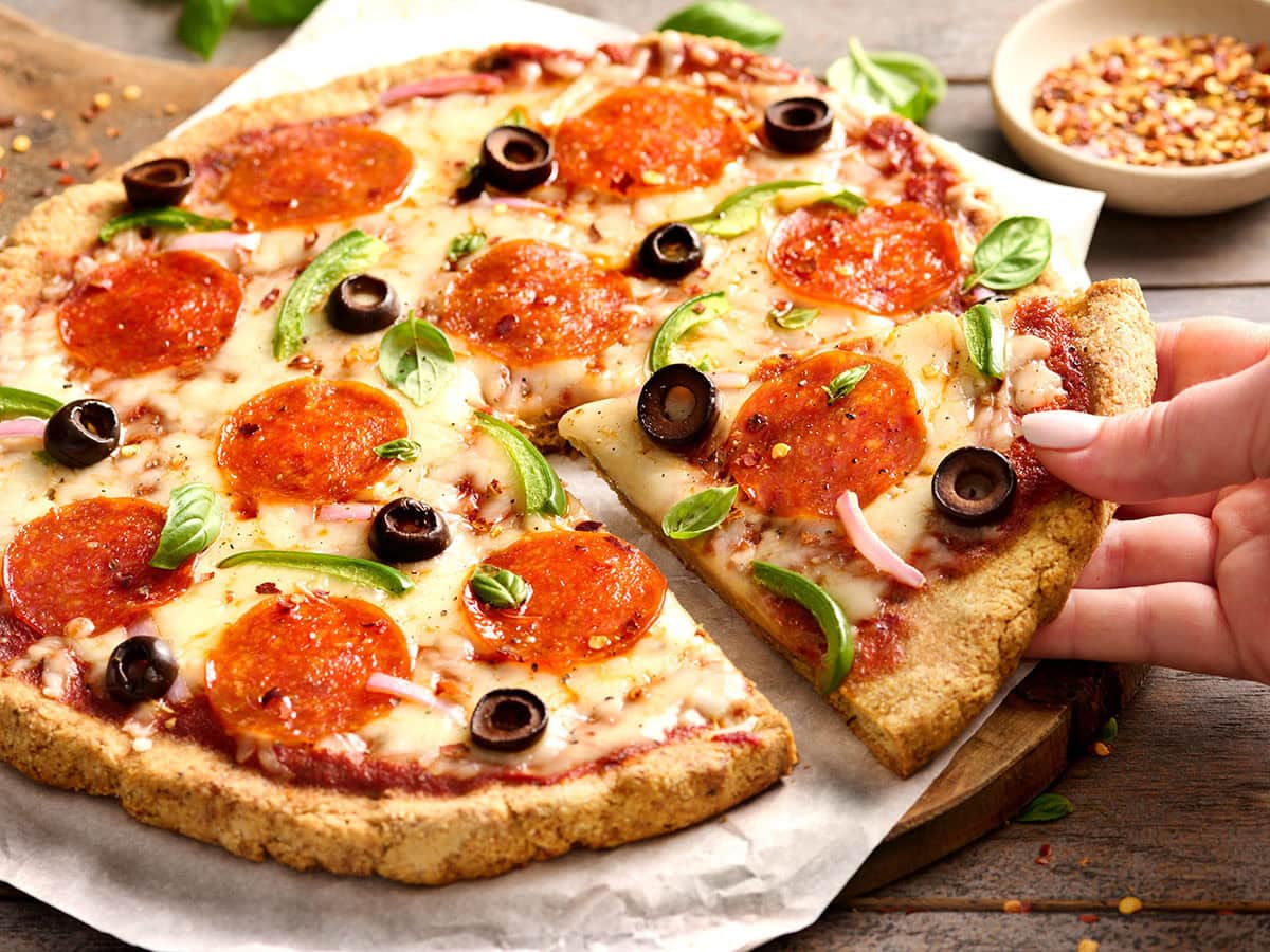 Hand lifting a slice of almond flour pepperoni pizza, garnished with fresh basil and red pepper flakes