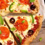 Close-up of almond flour pizza crust slices topped with pepperoni, black olives, green pepper, red onion, and fresh basil
