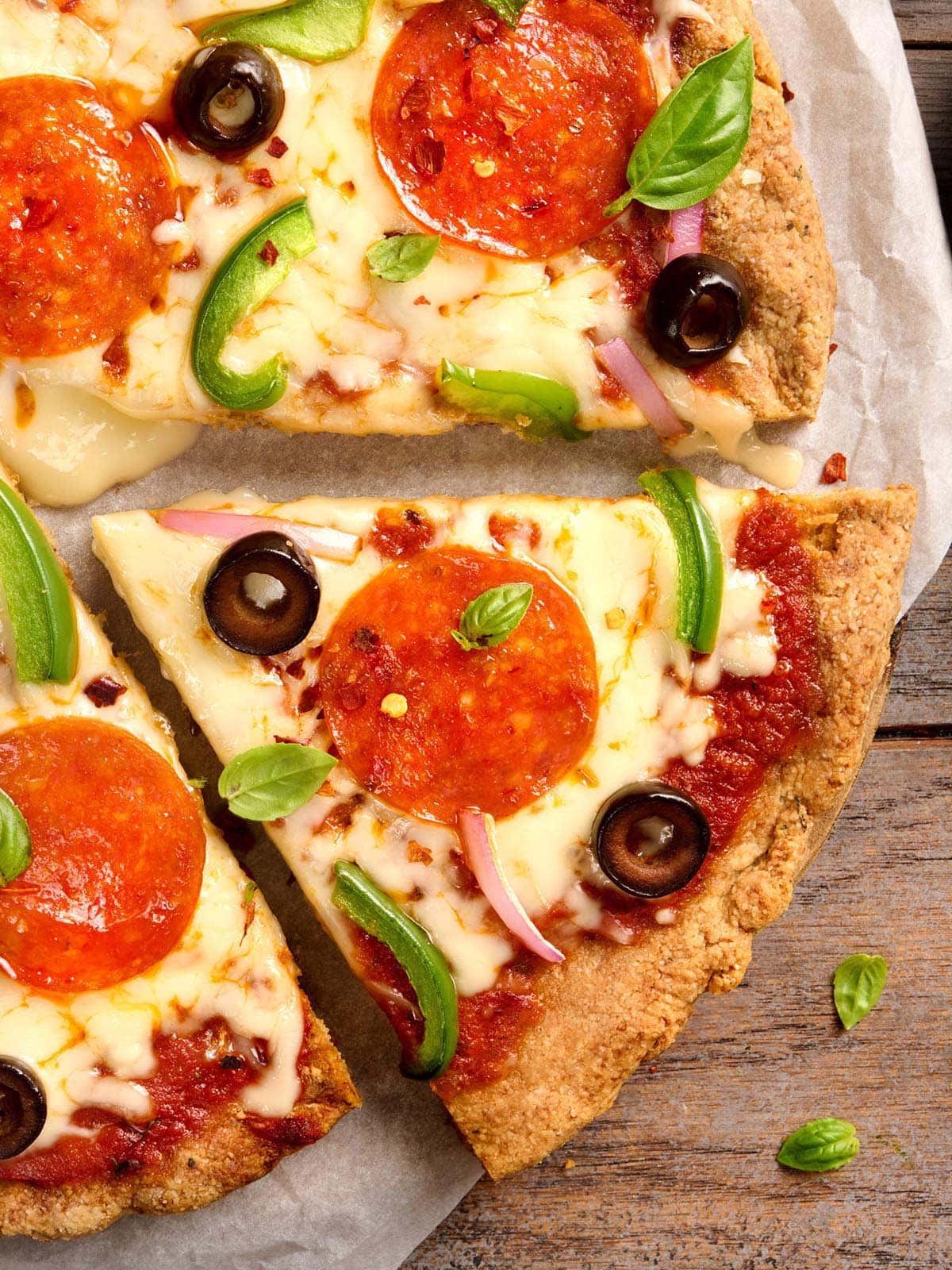 Close-up of almond flour pizza crust slices topped with pepperoni, black olives, green pepper, red onion, and fresh basil