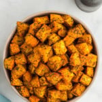 Bowl filled with golden roasted sweet potato cubes seasoned with herbs, placed on a light background with salt and pepper shakers nearby.