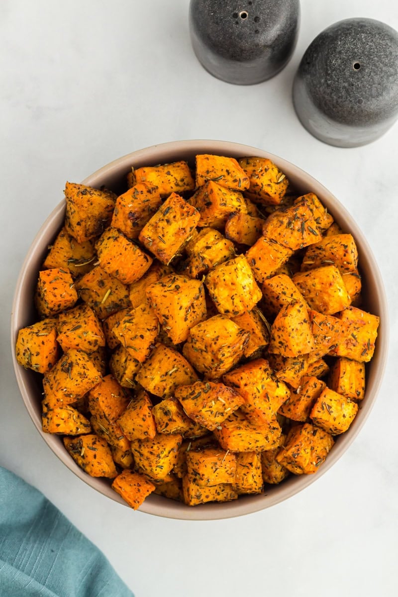 Bowl filled with golden roasted sweet potato cubes seasoned with herbs, placed on a light background with salt and pepper shakers nearby.