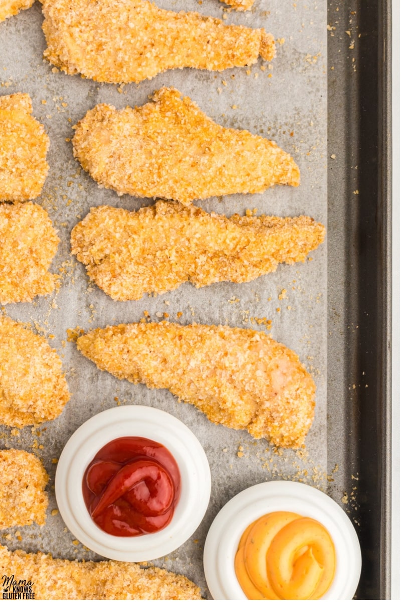 Gluten-Free Chicken Tenders and bowls of ketchup and mustard on parchment-lined baking tray.