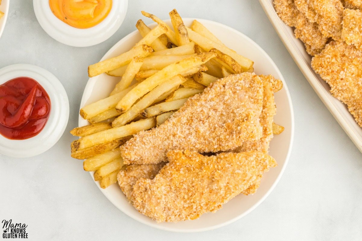 Gluten-Free Chicken Tenders and French fries on a round plate with ketchup and mustard in bowls on side.