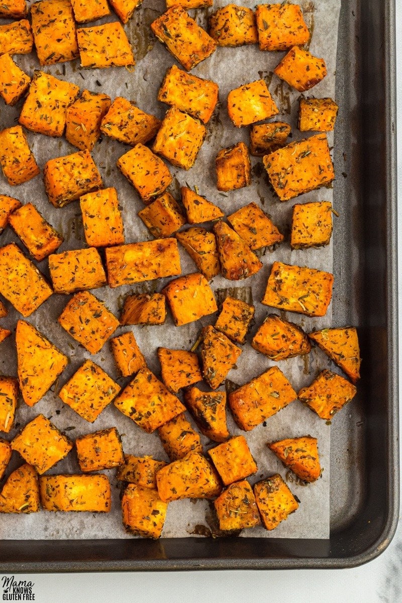 Roasted Sweet Potatoes cubes tossed in herbs on top of parchment-lined baking sheet.