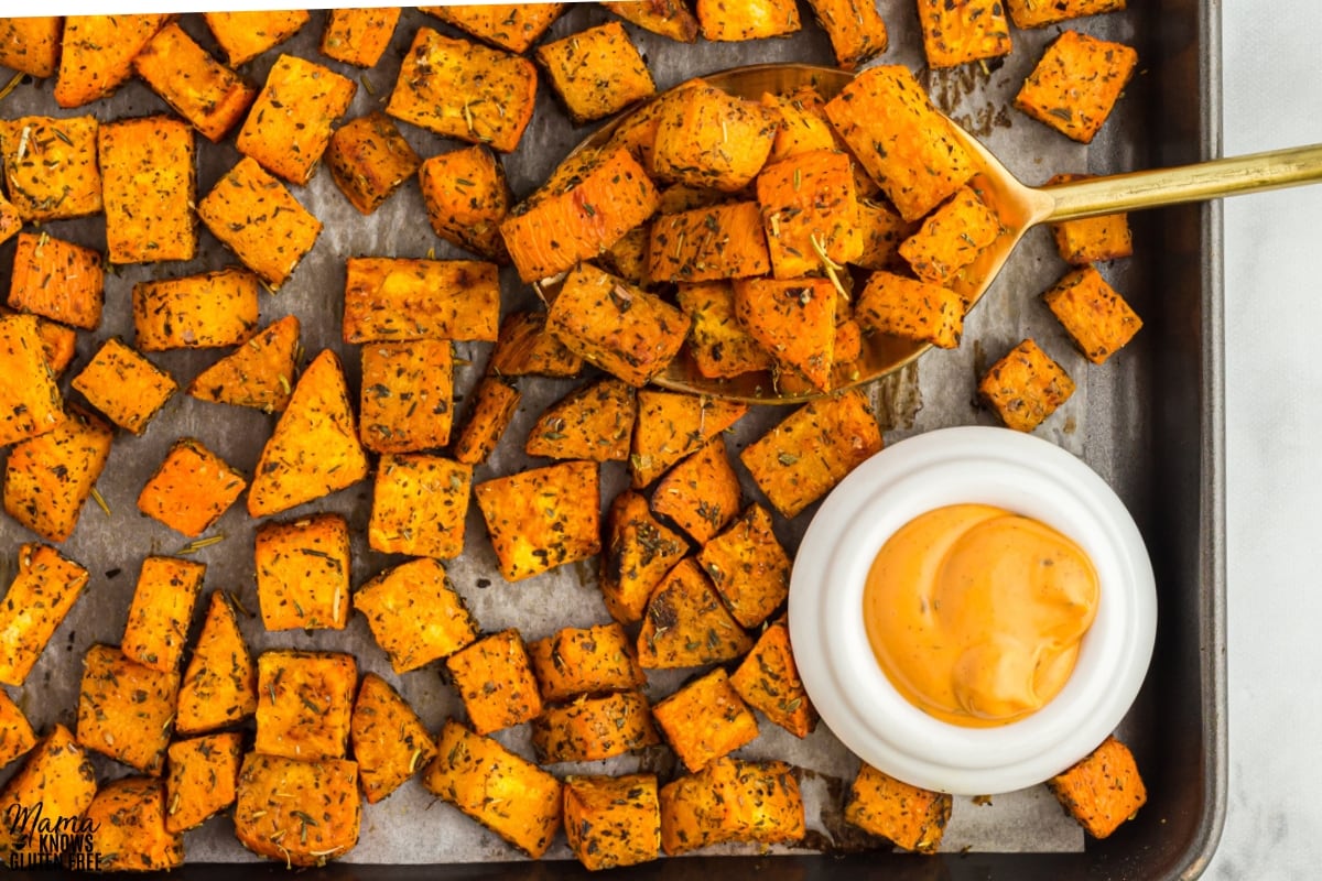 Roasted Sweet Potatoes cubes tossed in herbs on parchment-lined baking sheet with large spoon and small bowl of dipping sauce.