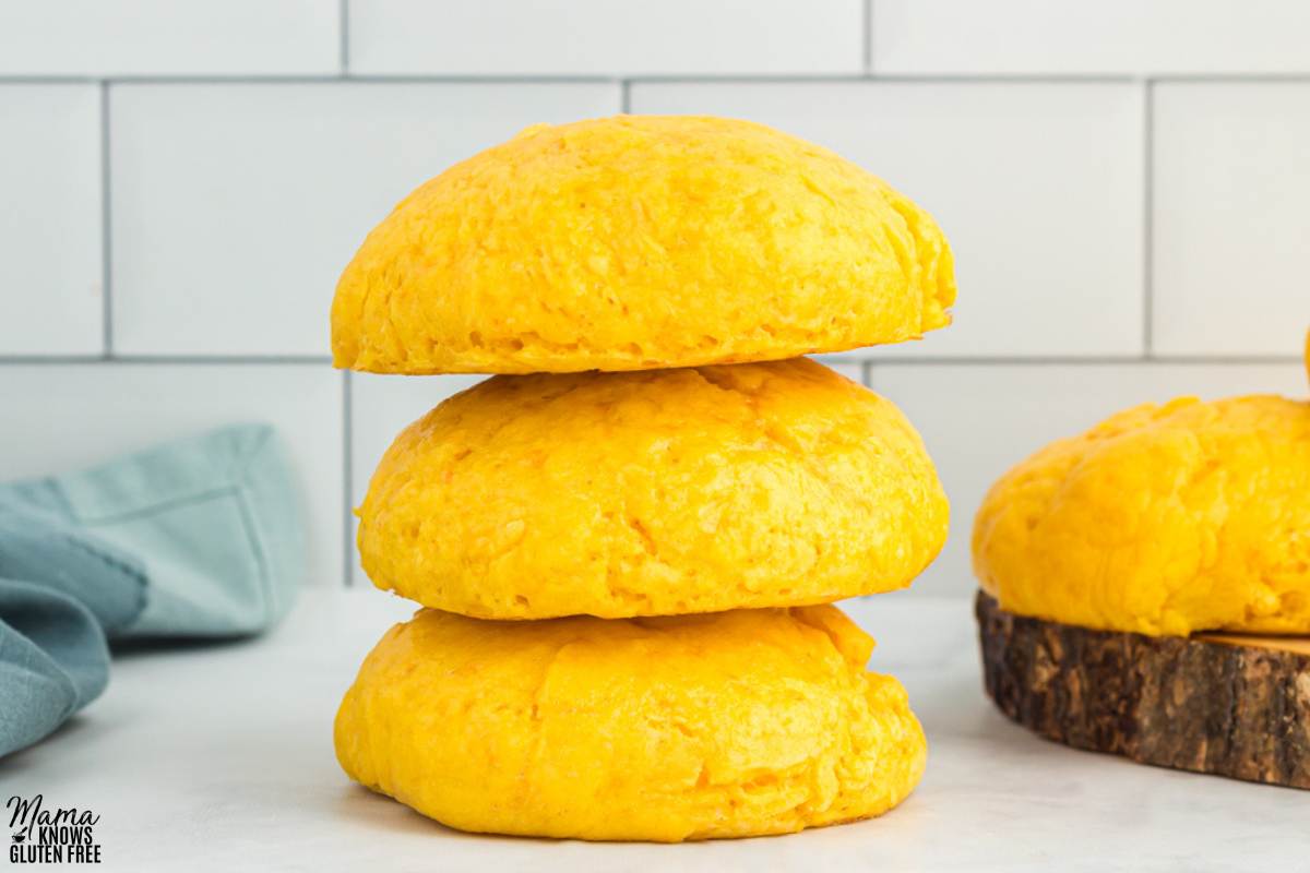 A pile of three Gluten-Free Sweet Potato Rolls on kitchen counter with towel and another roll in the background.