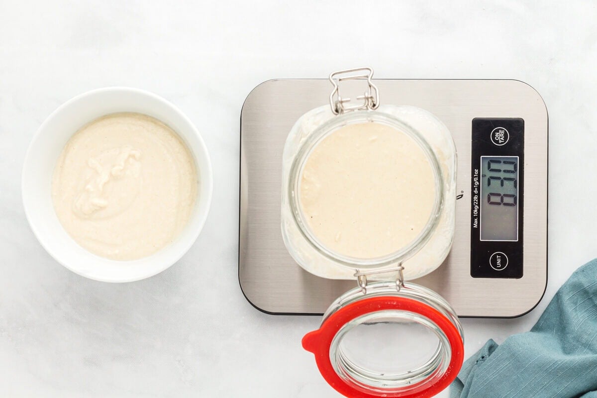 gluten free sourdough starter in a glass jar on a scale next to a bowl of starter discard.