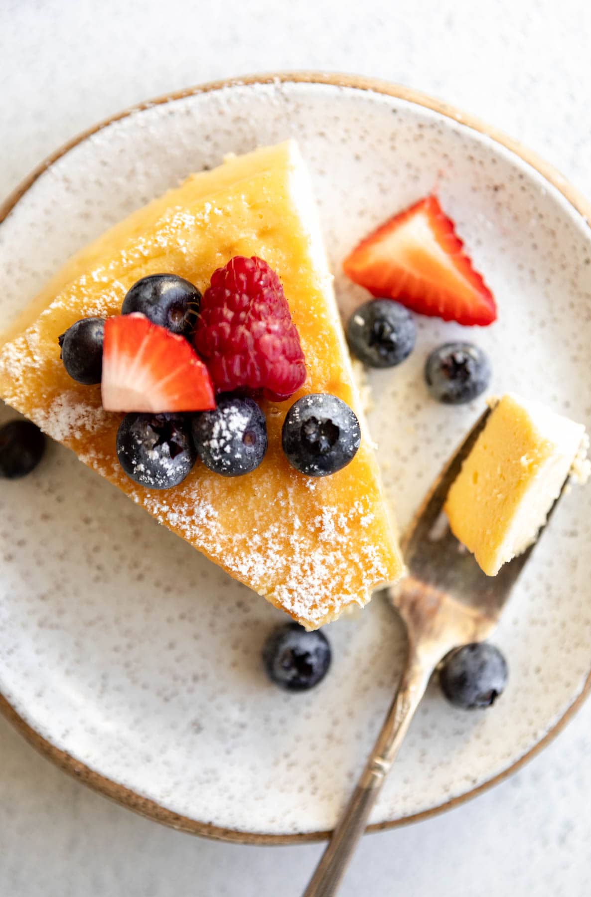 crustless cheesecake slice topped with fruit on a plate with a bite taken out by a fork.