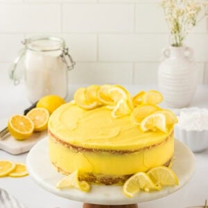 A frosted gluten-free lemon cake on a marble cake stand, decorated with lemon slices on top and around the base. The cake has a bright yellow frosting and a rustic, semi-naked finish. Fresh lemons, a jar of flour, and white ceramic vases are styled in the background.