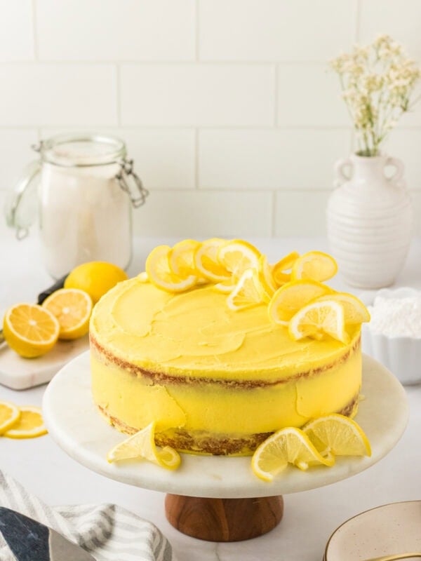 A frosted gluten-free lemon cake on a marble cake stand, decorated with lemon slices on top and around the base. The cake has a bright yellow frosting and a rustic, semi-naked finish. Fresh lemons, a jar of flour, and white ceramic vases are styled in the background.