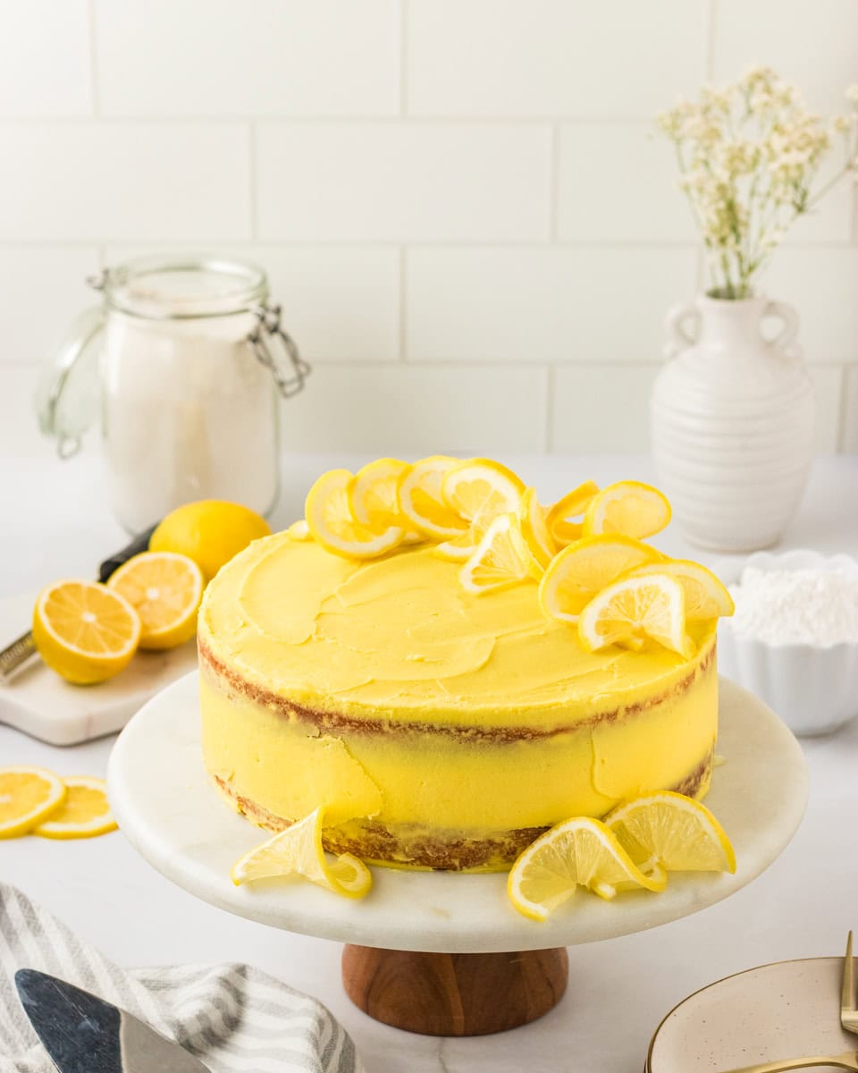 A frosted gluten-free lemon cake on a marble cake stand, decorated with lemon slices on top and around the base. The cake has a bright yellow frosting and a rustic, semi-naked finish. Fresh lemons, a jar of flour, and white ceramic vases are styled in the background.