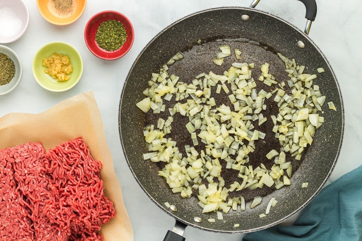 Sauteed onions in a skillet with raw ground beef next to it.