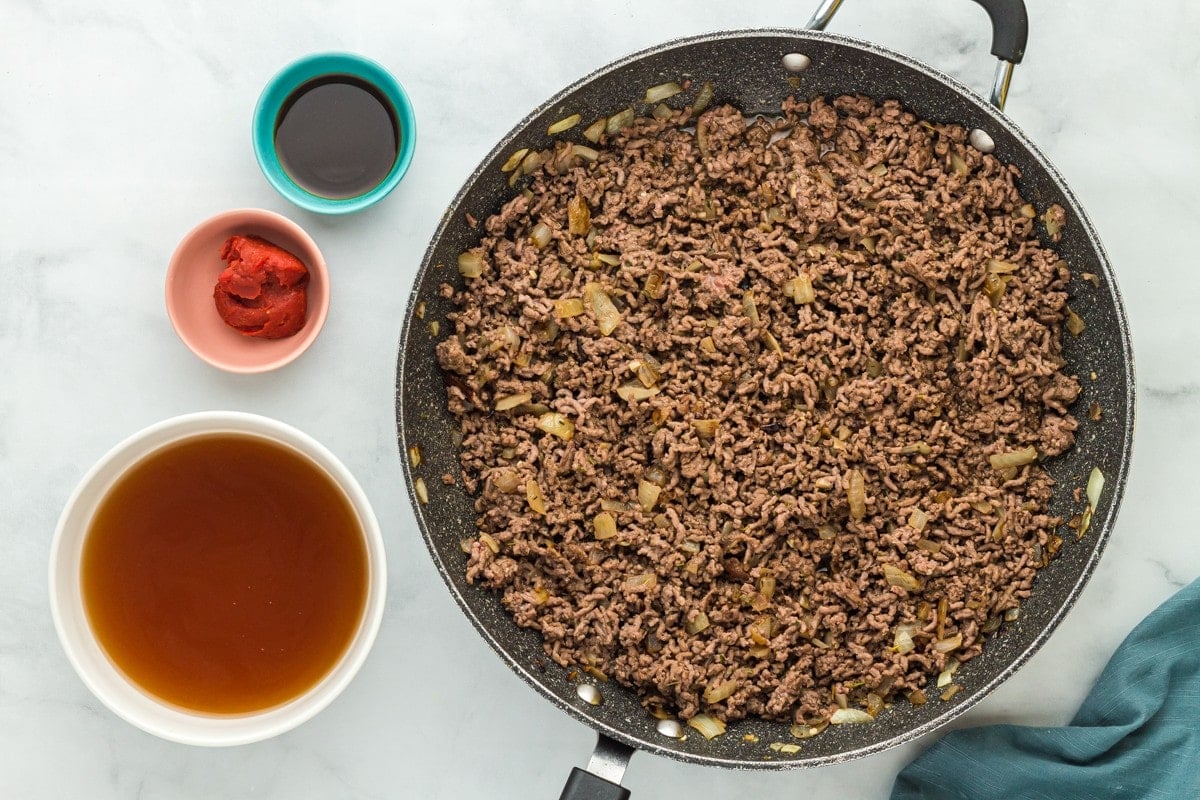 Cooked ground beef in a skillet with a bowl of broth, a bowl of tomato paste and a bowl of soy sauce next to it.