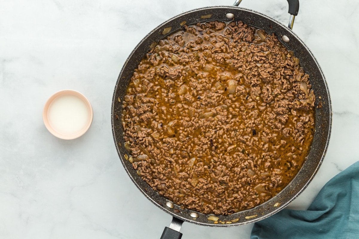 Seasoned ground beef in a skillet with a bowl of cornstarch and water next to it.