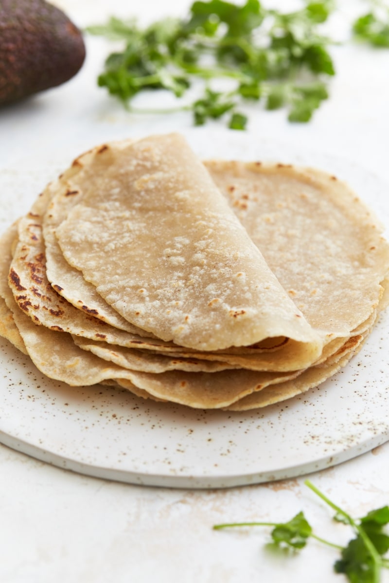 gluten-free flour tortillas folded on a white plate with an avocado and cilantro in the background