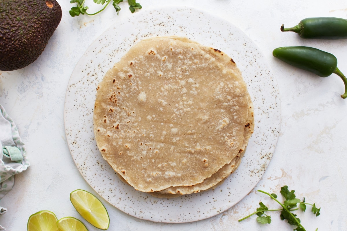 gluten-free flour tortillas on a white plate with an avocado, jalapenos, limes, and cilantro in the background