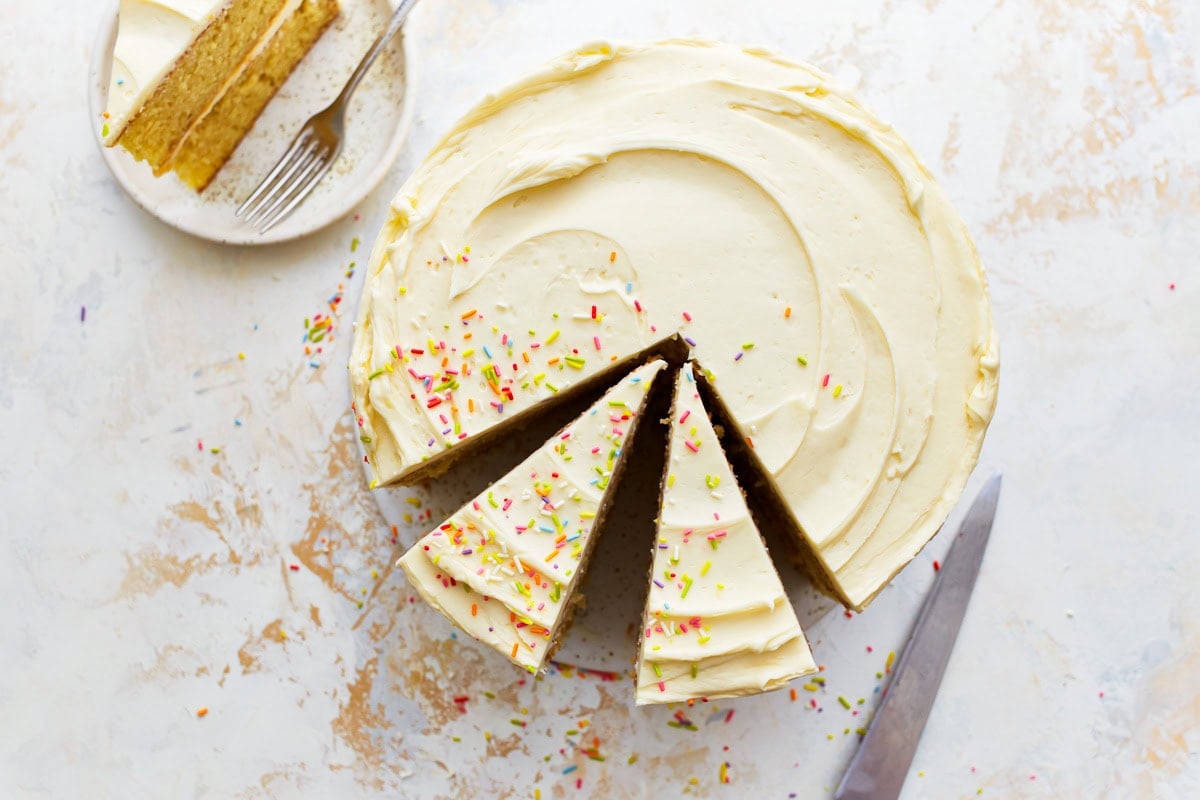 Top view of a round gluten-free vanilla cake frosted with vanilla buttercream and decorated with colorful sprinkles, with two slices cut and one slice plated with a fork.