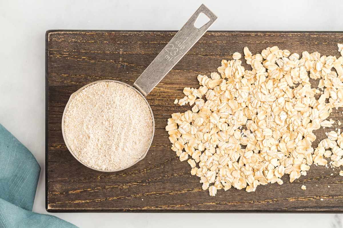 overhead view of oat flour in a measuring cup on a wooden cutting board with whole oats.