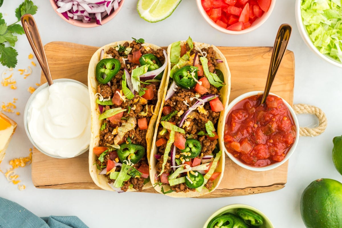 overhead view of 2 gluten-free tacos on a wooden cutting board with taco sauce and sour cream.