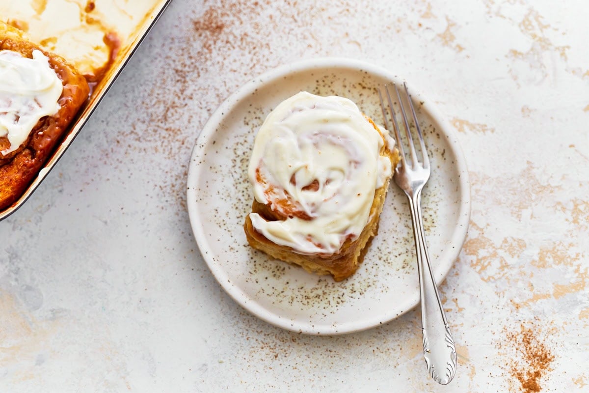 A frosted gluten-free cinnamon roll on a speckled ceramic plate with a fork beside it.