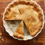 overhead view of a gluten-free chicken pot pie in a white pie pan on a wooden countertop with 2 slices gone and another slice cut