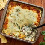 overhead shot of a spoon dipped into a tin square baking dish with hot spinach dip topped with mozzarella cheese on a wooden surface