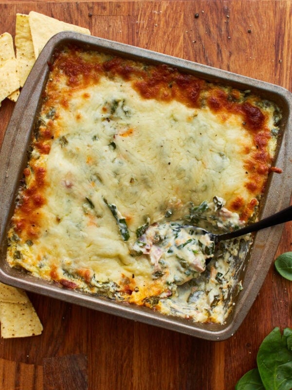 overhead shot of a spoon dipped into a tin square baking dish with hot spinach dip topped with mozzarella cheese on a wooden surface
