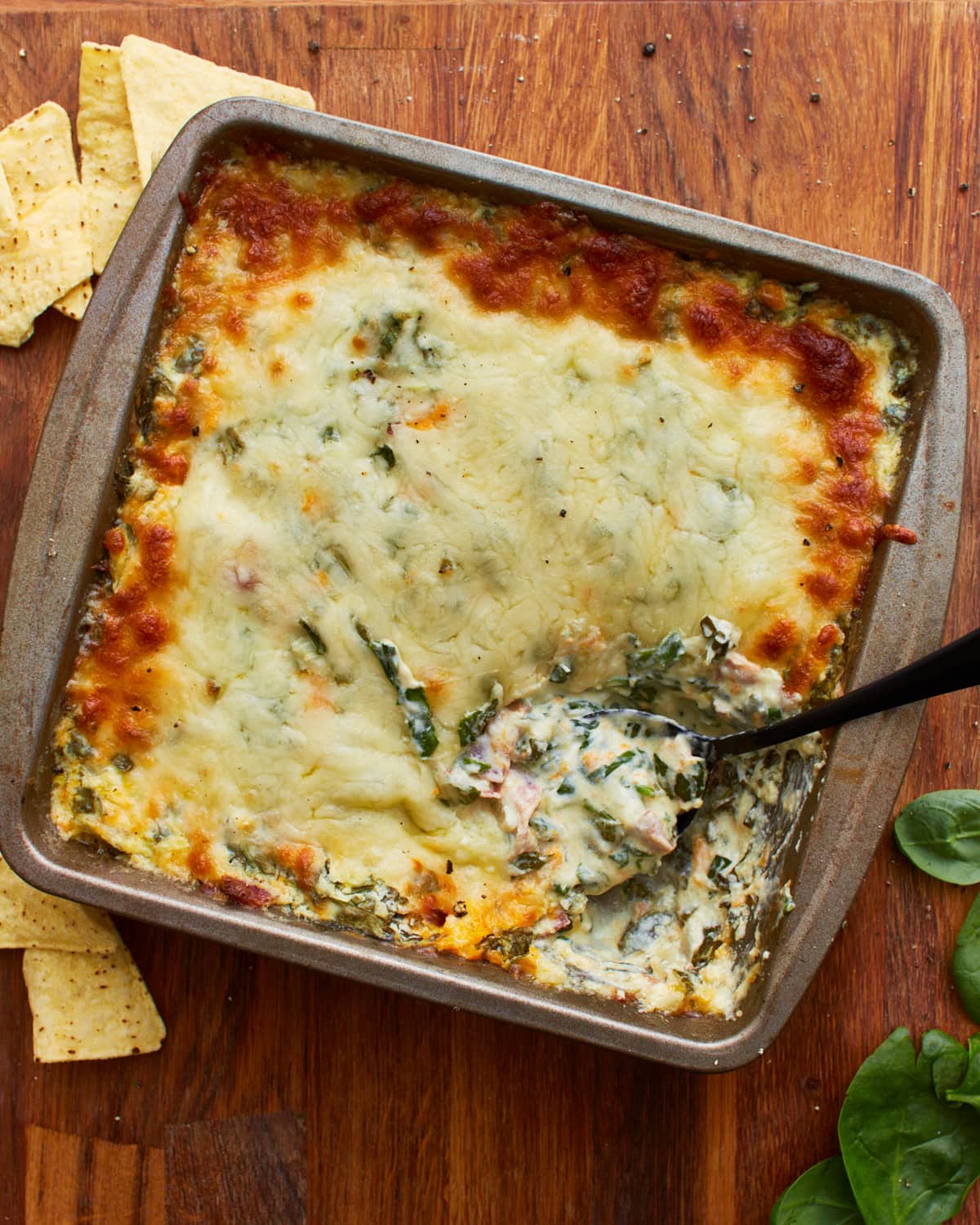 overhead shot of a spoon dipped into a tin square baking dish with hot spinach dip topped with mozzarella cheese on a wooden surface