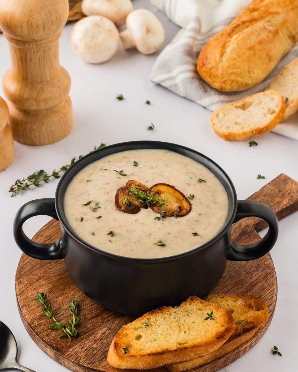 A creamy bowl of gluten-free cream of mushroom soup served in a black soup bowl, garnished with thyme and roasted mushroom slices. The bowl sits on a wooden coaster, accompanied by toasted baguette slices. Mushrooms, bread, and thyme sprigs are scattered in the background