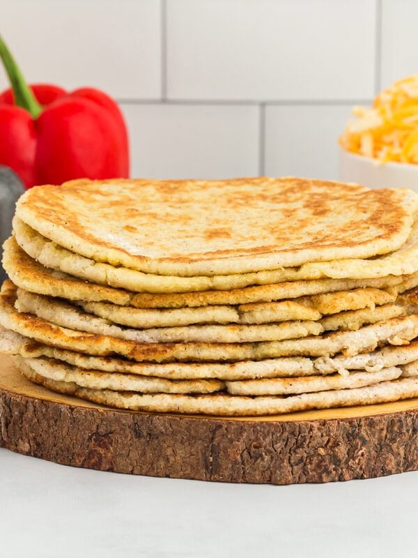 stacked coconut flour tortillas on a wooden board with bell peppers in the back and shredded cheese in a white bowl