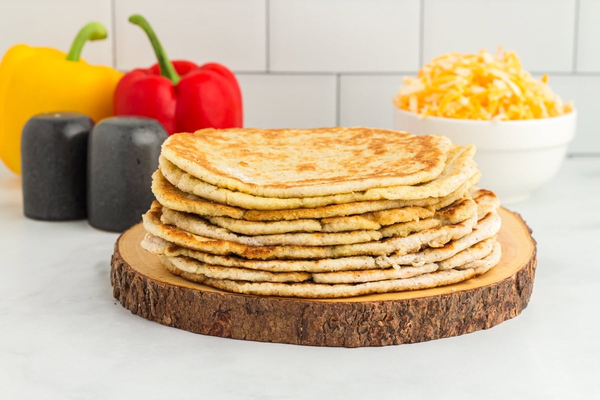stacked coconut flour tortillas on a wooden board with bell peppers in the back and shredded cheese in a white bowl