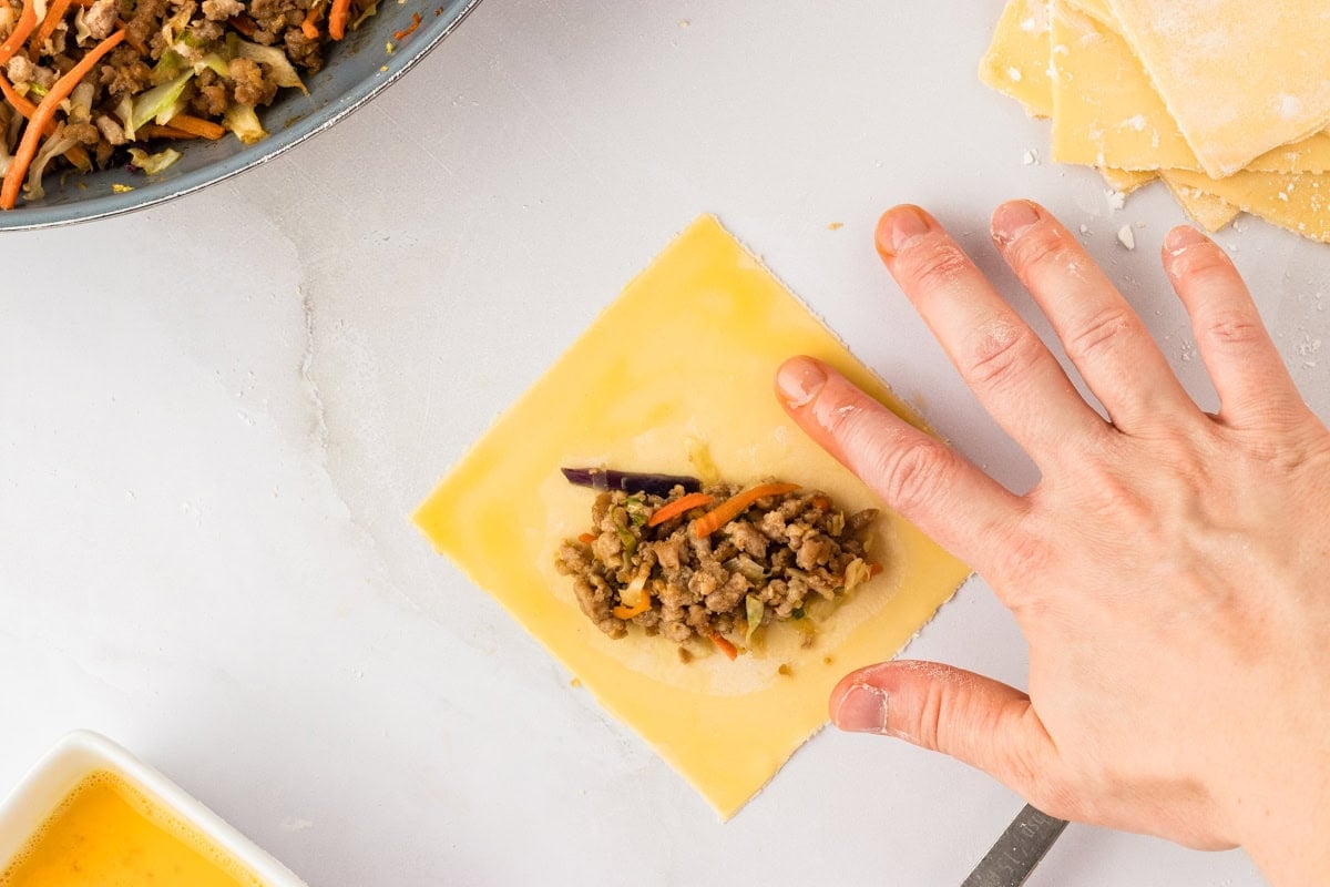 hand putting egg wash on edges of gluten-free egg roll dough square with filling in the center