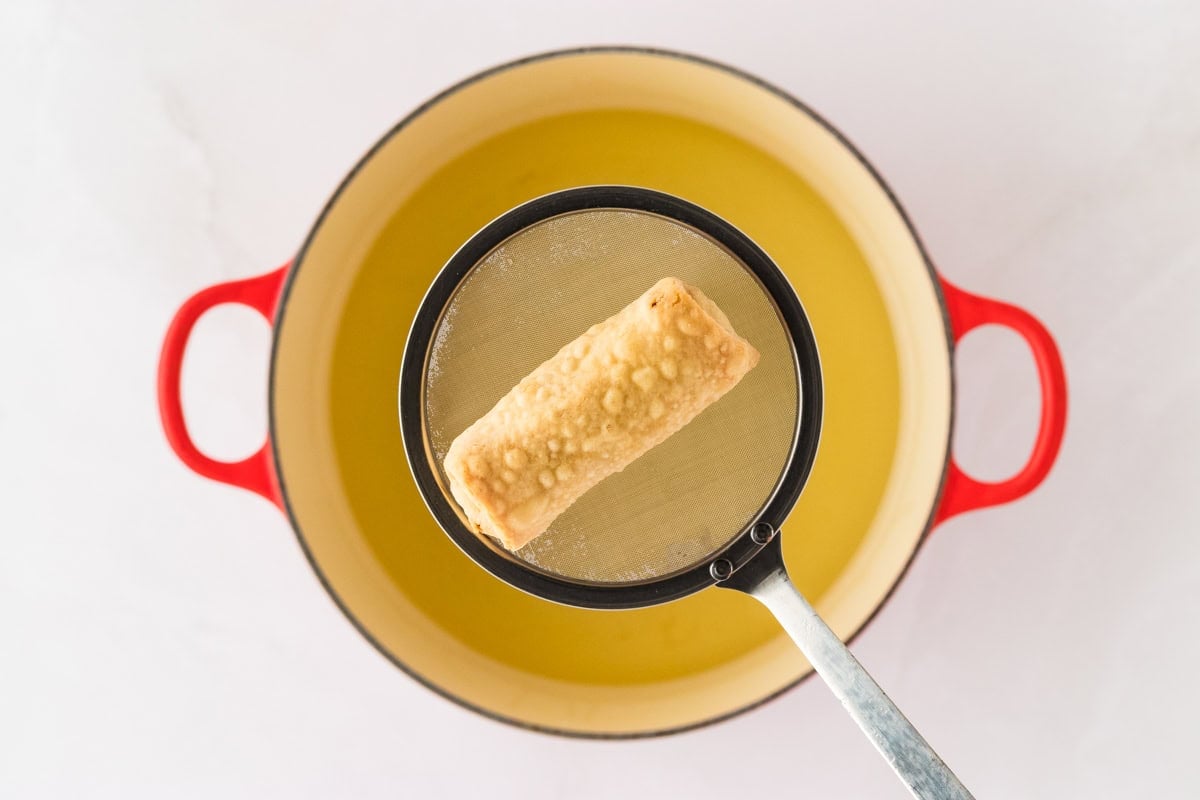 1 cooked gluten-free egg roll in skimmer over red pot with frying oil