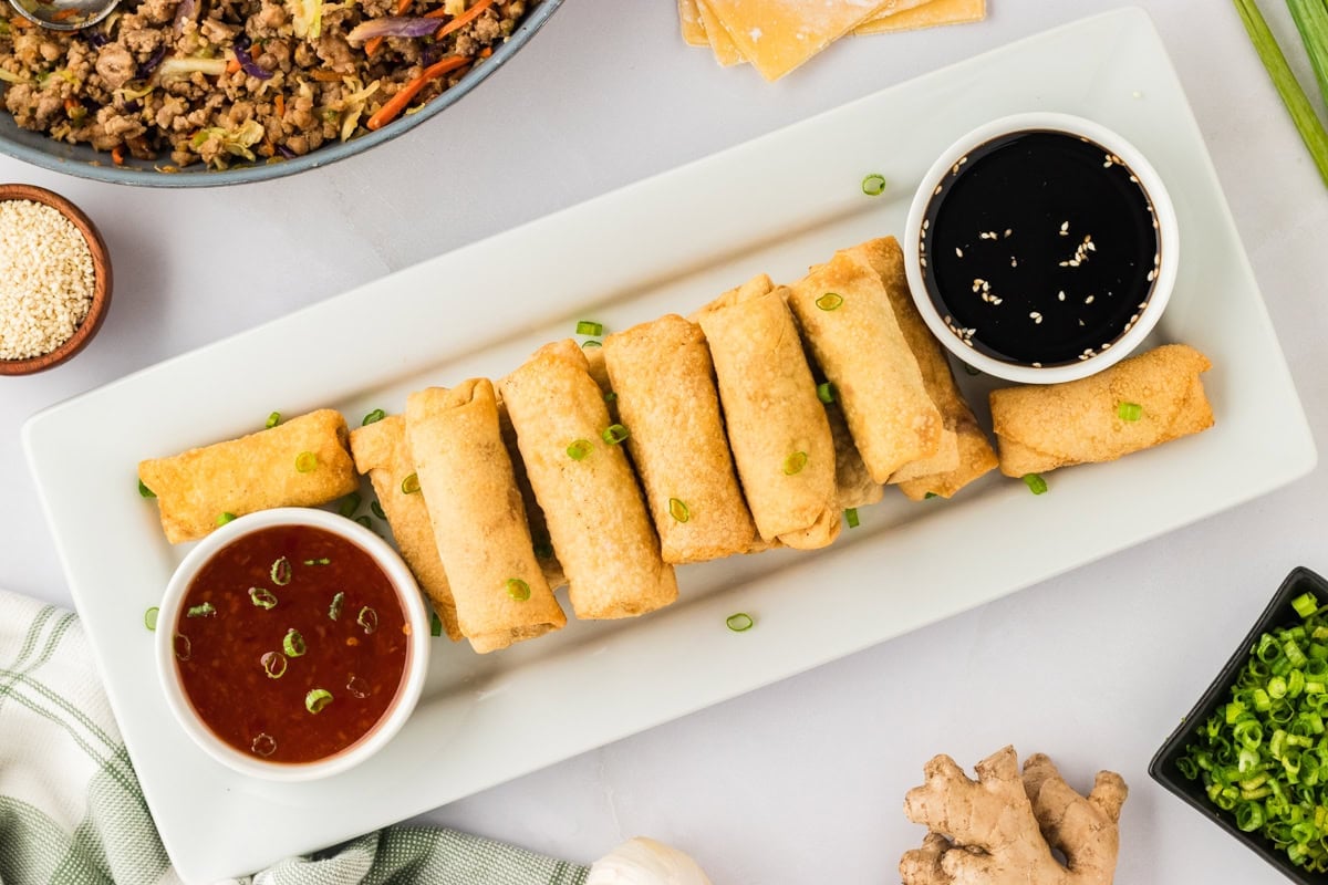 a bunch of fried gluten-free egg rolls on a white rectangular plate with sweet chili sauce and soy sauce in small white bowls