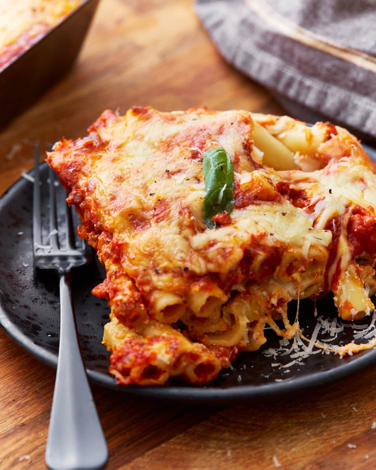 front on view of gluten-free baked ziti on black plate with a leaf of fresh basil on top and a black fork