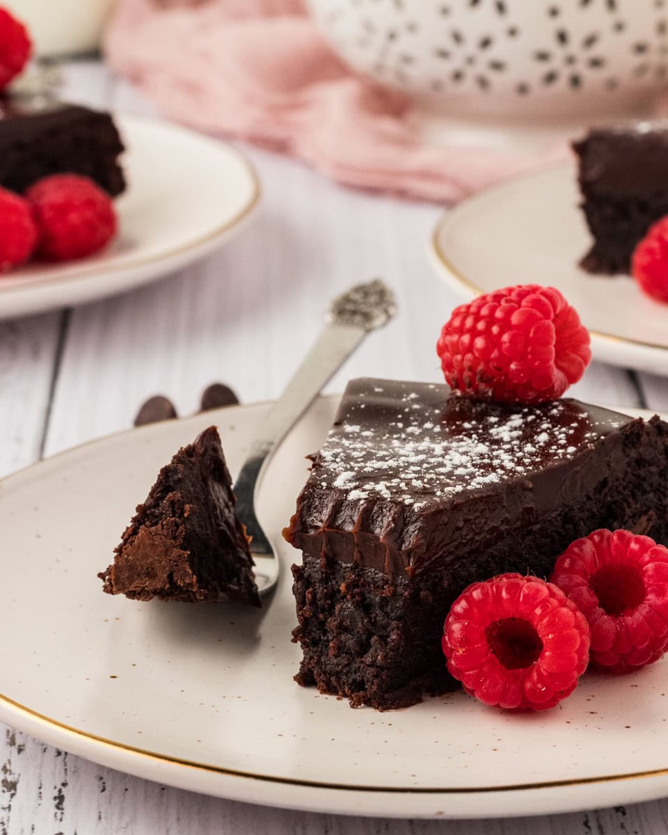flourless chocolate cake piece on white plate with powder sugar, raspberries and a piece taken out on a silver spoon