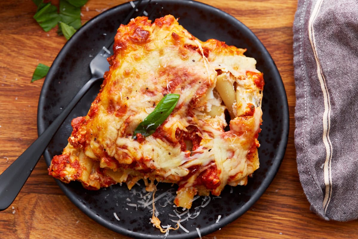 piece of gluten-free baked ziti on a black plate with a black fork next to a gray rag