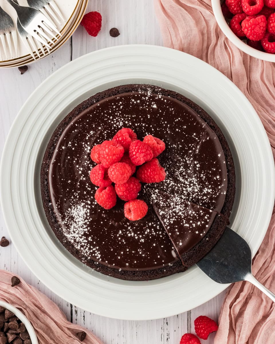 Metal cake knife lifting up piece of flourless chocolate cake from the whole cake, topped with powdered sugar and raspberries on a white plate