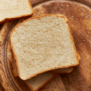 Two pieces of gluten-free bread stacked on a round wooden cutting board with another piece of bread in the background