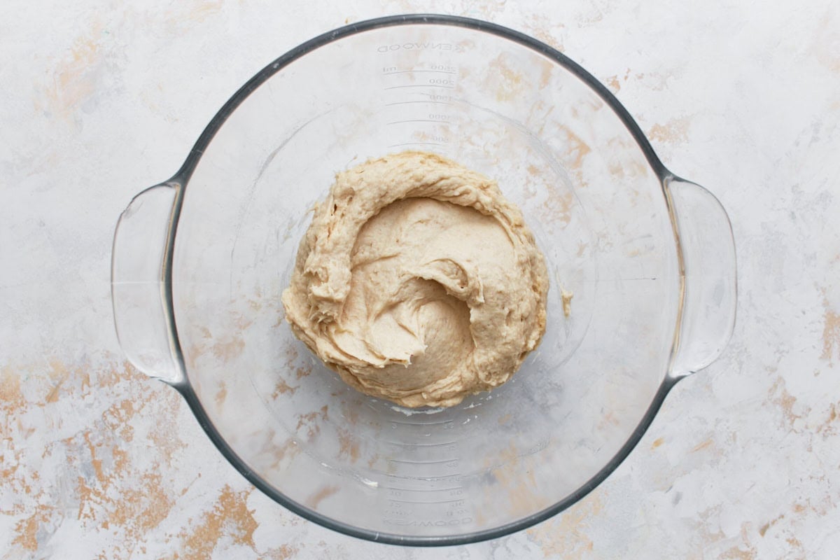 A glass mixing bowl containing a smooth, light beige gluten-free bread dough after being mixed, placed on a light-colored countertop