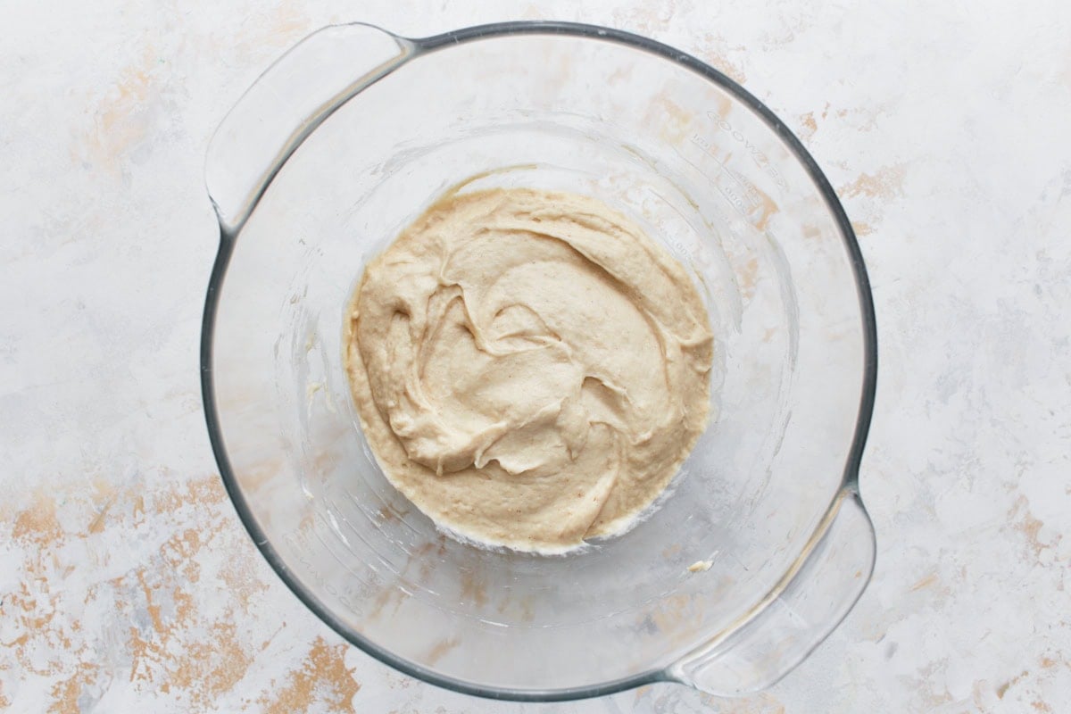 A glass mixing bowl containing a smooth, light beige gluten-free bread dough after being mixed, placed on a light-colored countertop
