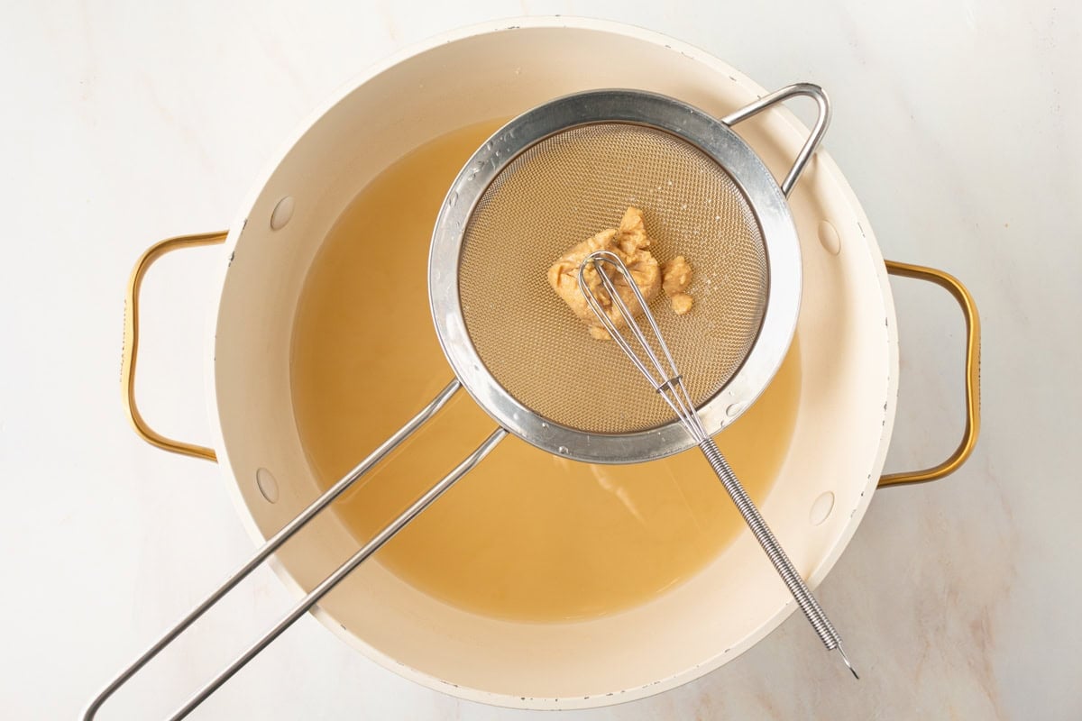 a small sieve with white miso paste being whisked into gluten-free miso soup in a large pot
